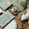 Luxury sage green wedding invitation suite styled with floral details cards, vintage postage, custom envelope liner artwork, wax seal accents, elegant menu card, bridal shoes, and wedding rings on a rustic wood surface.