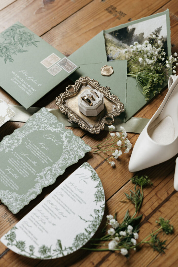 Luxury sage green wedding invitation suite styled with floral details cards, vintage postage, custom envelope liner artwork, wax seal accents, elegant menu card, bridal shoes, and wedding rings on a rustic wood surface.