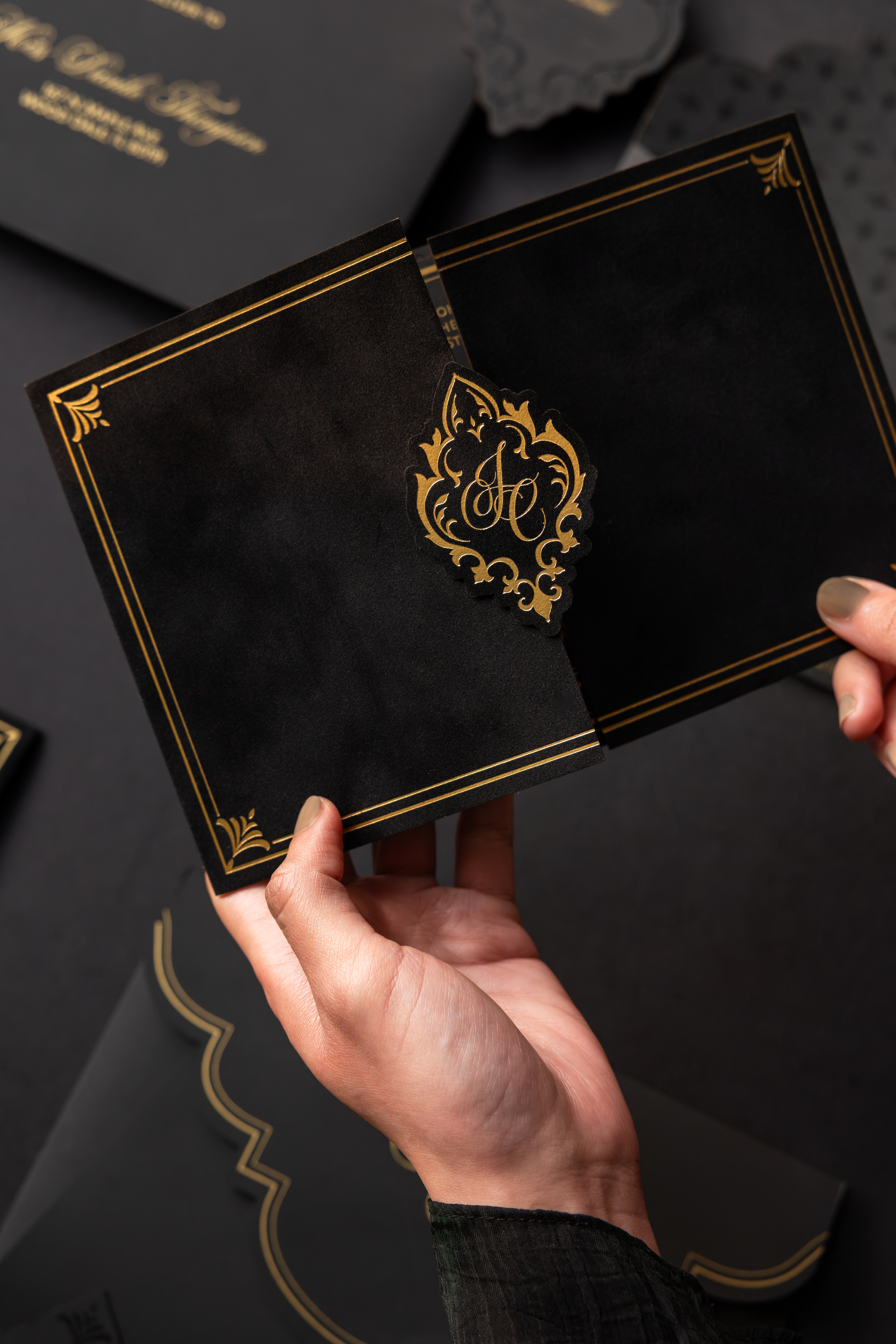 Hands opening a luxurious black wedding invitation folio with ornate gold foil monogram detailing and elegant corner accents from a sophisticated black-tie wedding suite.