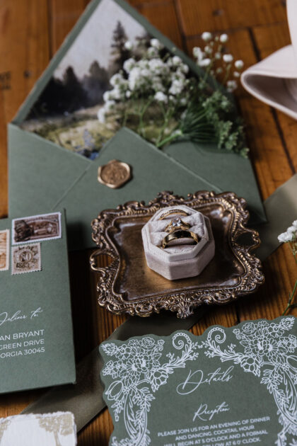 Elegant sage green wedding invitation details featuring floral illustrated stationery, vintage postage, wax seal envelope, wedding rings styled on an ornate tray, and romantic garden-inspired wedding details on a rustic wood background.