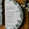 Elegant half-moon wedding menu card featuring botanical floral illustrations and bird details in deep green ink on textured white cardstock, styled on a rustic wood surface.