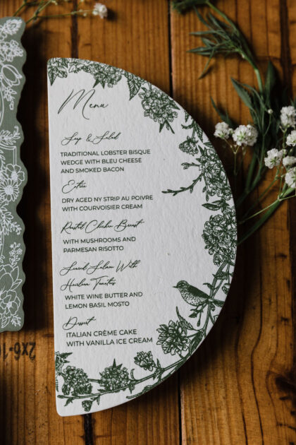 Elegant half-moon wedding menu card featuring botanical floral illustrations and bird details in deep green ink on textured white cardstock, styled on a rustic wood surface.