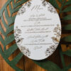 Elegant oval wedding menu card featuring botanical floral and bird illustrations in deep green ink on textured white cardstock, styled atop a dark green laser-cut charger on a rustic wood table.