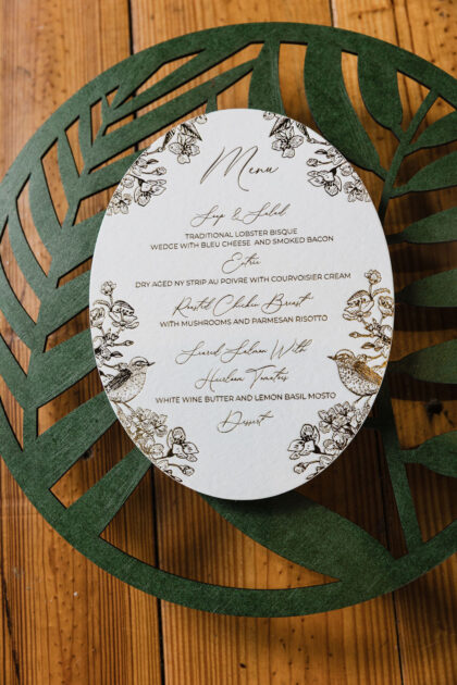 Elegant oval wedding menu card featuring botanical floral and bird illustrations in deep green ink on textured white cardstock, styled atop a dark green laser-cut charger on a rustic wood table.