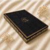 Luxury black lace wedding invitation box with intricate laser-cut detailing, gold foil monogram, and ornate gold foil edging on an ivory fabric backdrop.